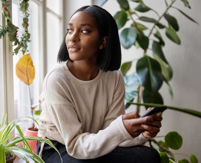 Kvinna som tittar ut genom ett fönster med en mobiltelefon i handen